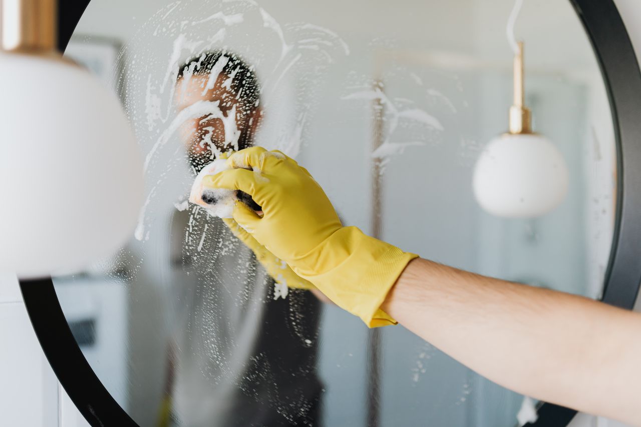 Services-03 Anonymous male in yellow rubber gloves wiping foam from round mirror in bathroom